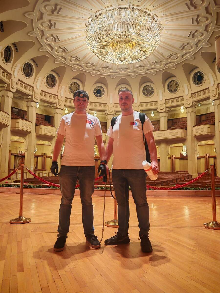 Doi operatori Compania DDD la o intervenție într-o sală de spectacole, cu pulverizator cu lance, rezervor purtat în spate, mănuși și lanternă frontală